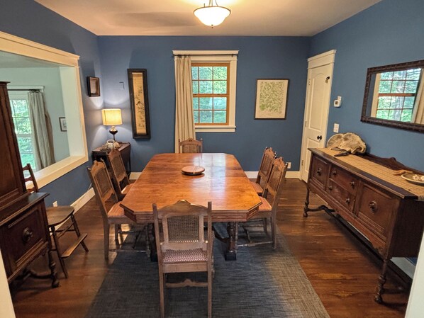 A dining room with a wooden table, chairs, a chandelier, and a view of the outdoors through windows.