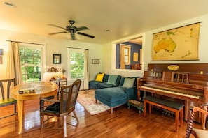 A cozy living room with a wooden floor, a dining table with chairs, a blue sofa, a piano, and a ceiling fan.