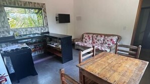 Un coin repas avec une table et des chaises en bois, un canapé recouvert d’un tissu à motifs floraux, un téléviseur fixé au mur et un espace cuisine avec un dosseret carrelé.