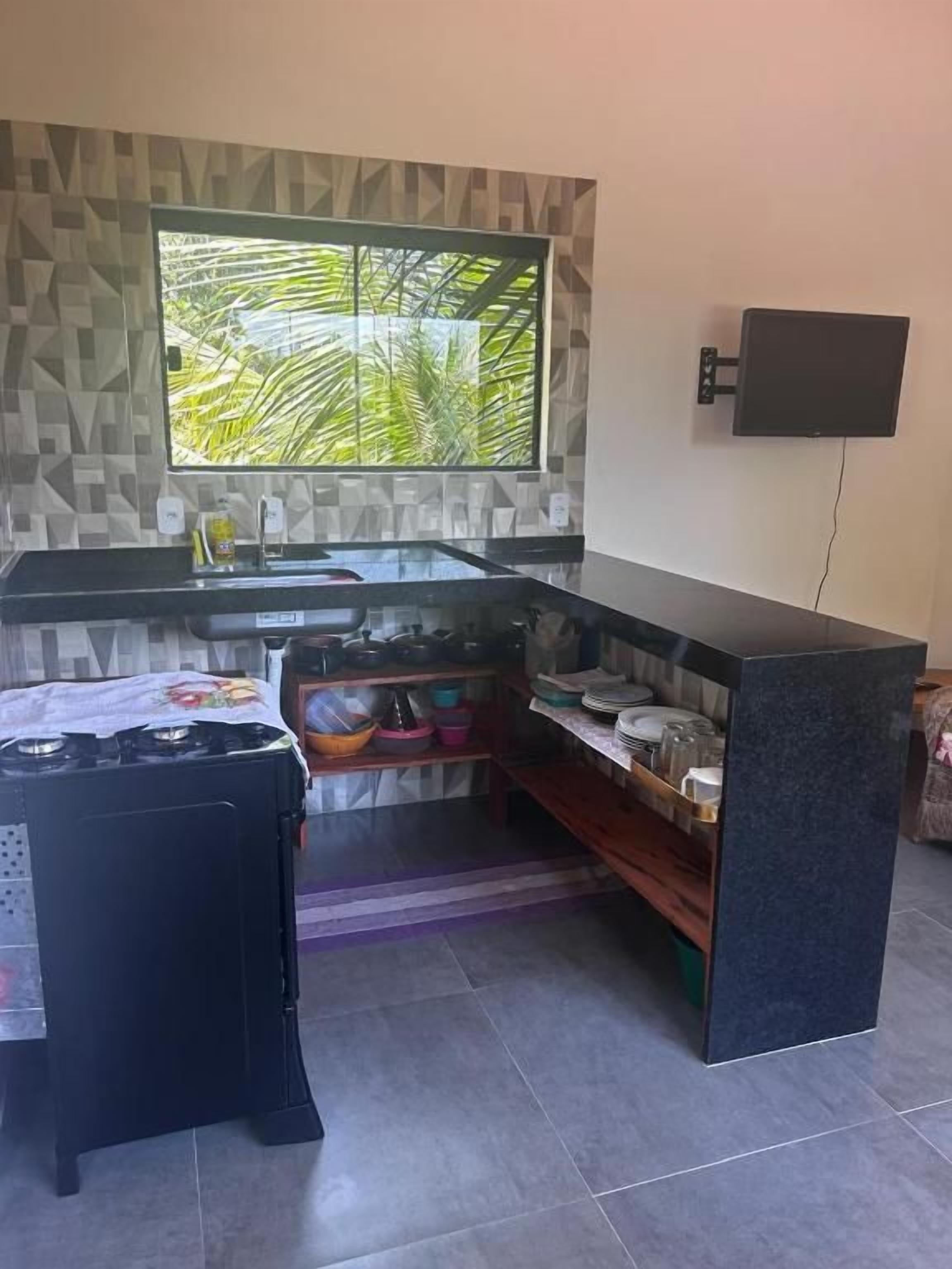 A compact kitchen with a built-in stove, a window with a view of palm trees, and a flat-screen TV mounted on the wall.