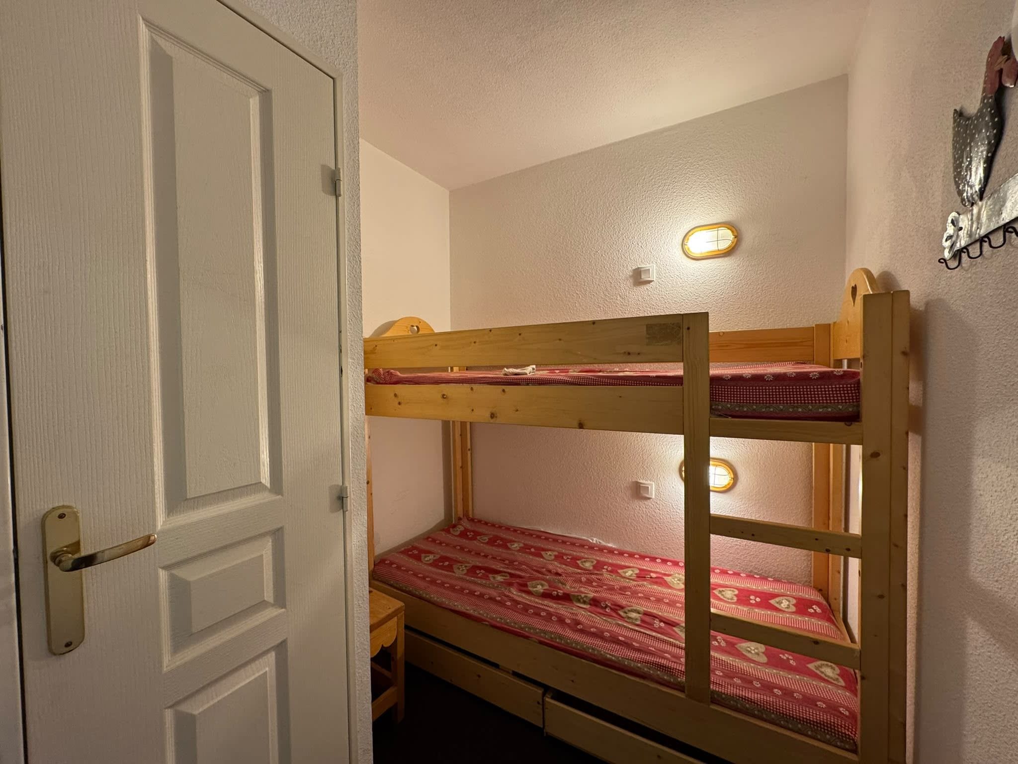 A bunk bed with a red patterned bedspread, a small wooden desk, and a wall-mounted light fixture.