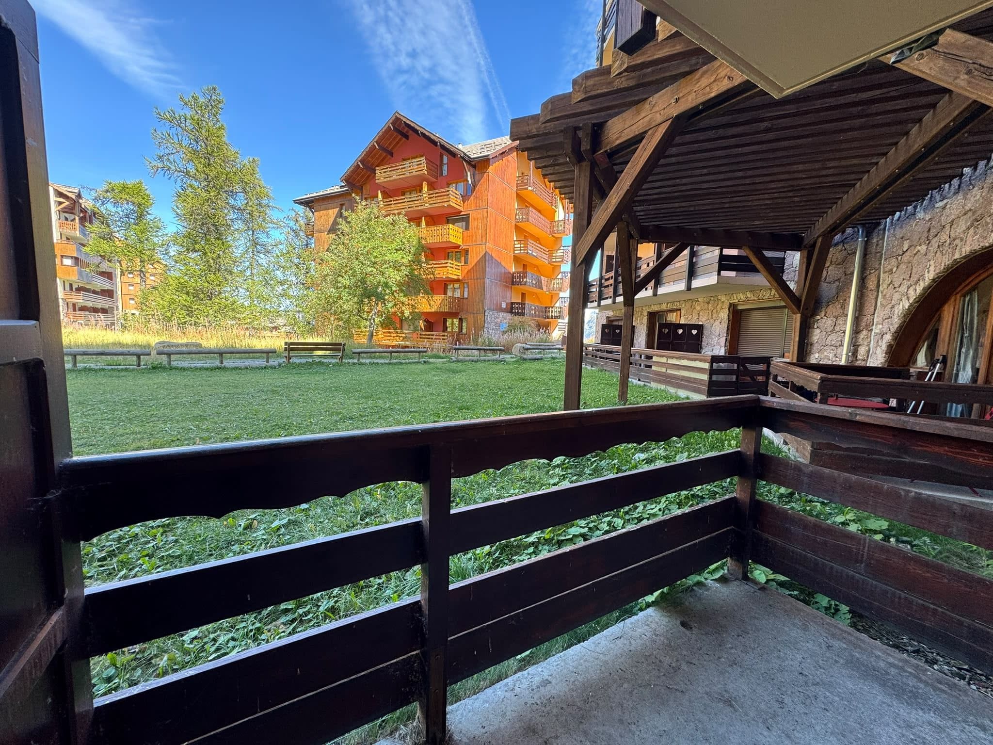 A view from a balcony with a wooden railing, overlooking a grassy area and a multi-story building with balconies.