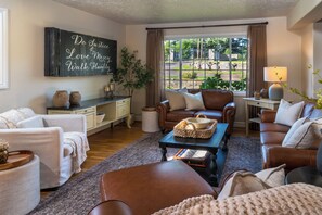 A cozy living room with a fireplace, comfortable seating, and a view of a garden through large windows. - 7BR Family Retreat + Private Pond+ Outdoor Kitchen (Oregon City)