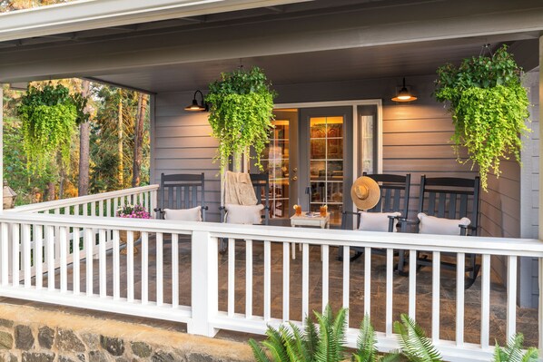 A porch with a white railing, two chairs, a table, hanging plants, and a door with glass panels. - 7BR Family Retreat + Private Pond+ Outdoor Kitchen (Oregon City)