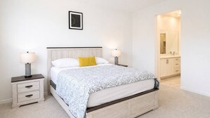A bedroom with a bed, bedside tables, a nightstand, a framed picture on the wall, and a bathroom visible through an open door. - Fully Furnished 3BR and 3BA Retreat with Views (Eastvale)