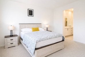 A bedroom with a bed, bedside tables, a nightstand, a framed picture on the wall, and a bathroom visible through an open door.