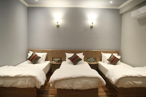 Ein Hotelzimmer mit drei Einzelbetten, jeweils mit einem Holzkopfteil, weißer Bettwäsche und dekorativen Kissen. Es gibt Nachttische mit Lampen und eine wandmontierte Klimaanlage.