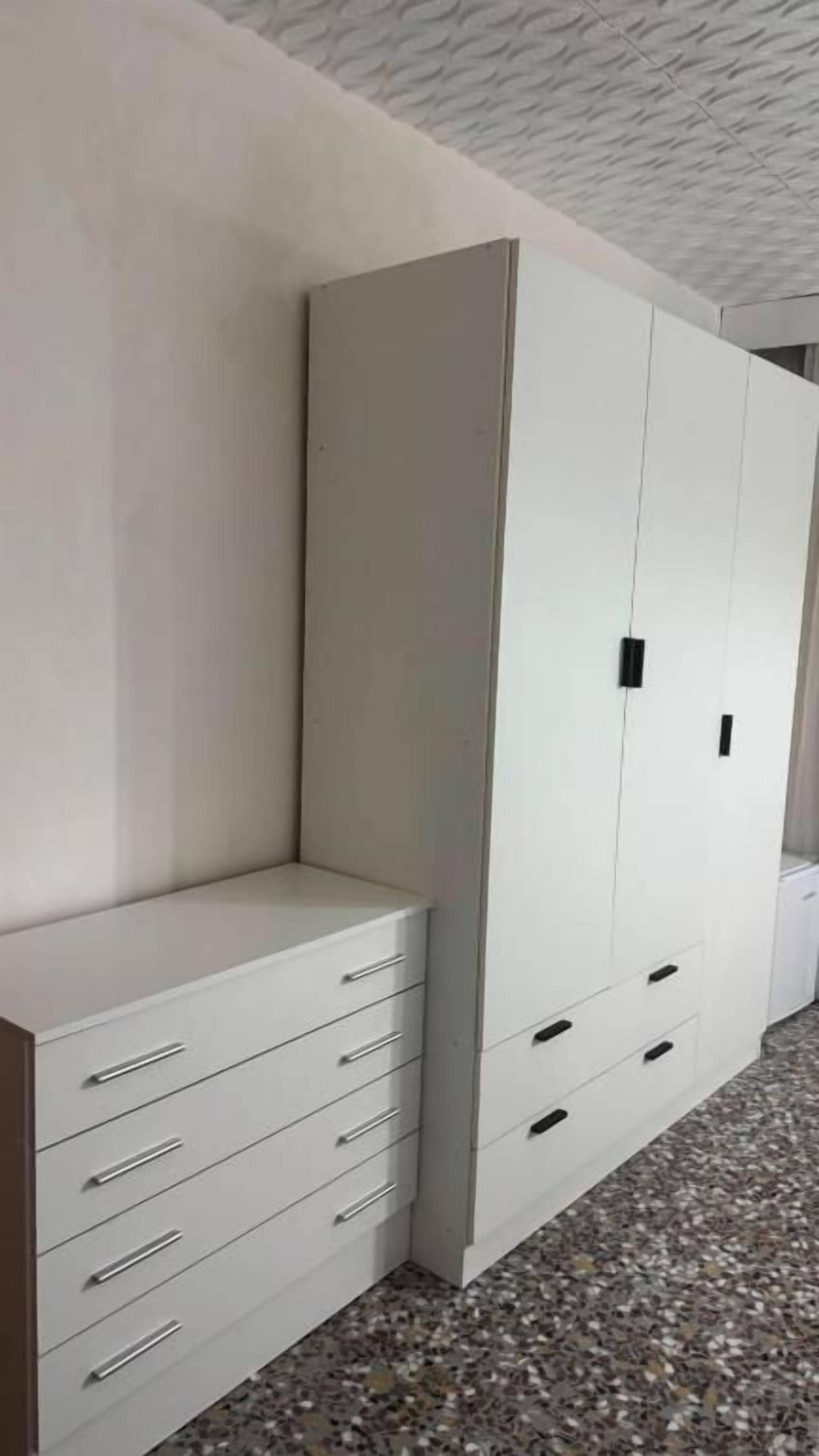 Quarto com guarda-roupa branco, penteadeira branca e chão revestido de azulejos.