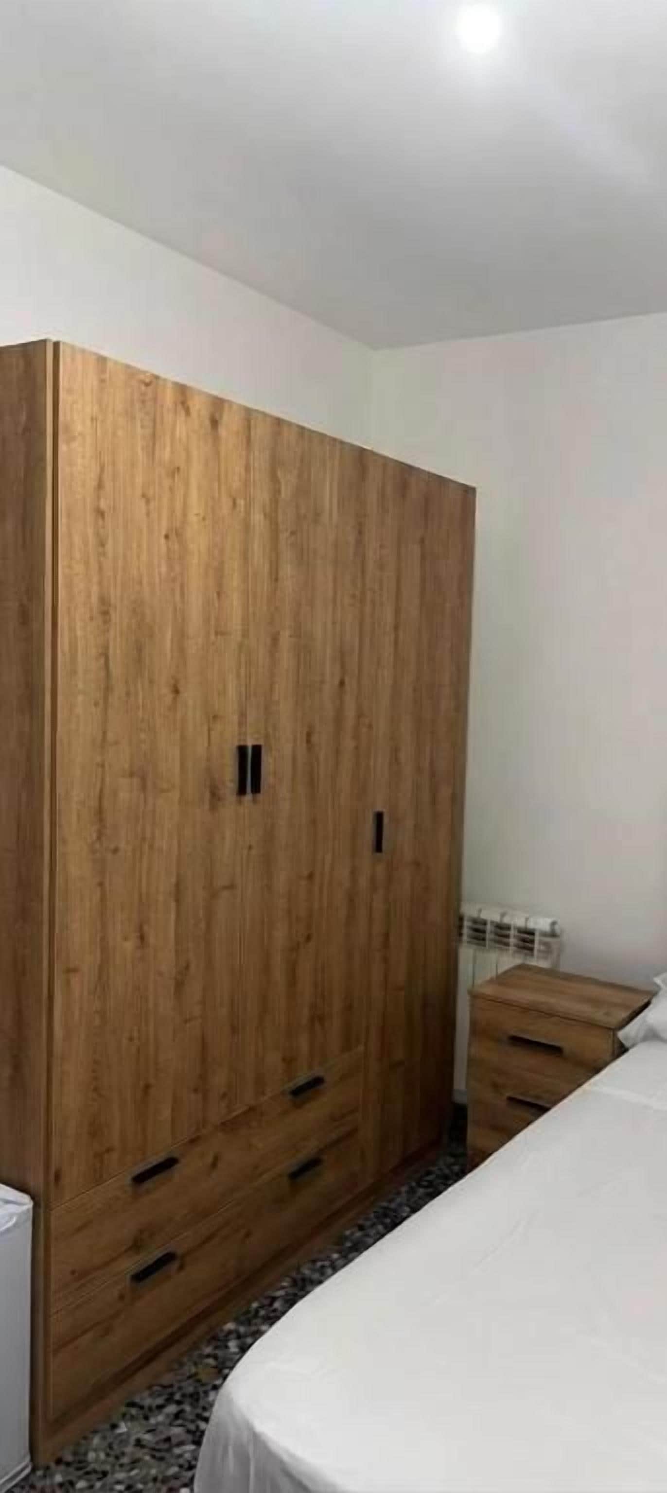 Un dormitorio con un amplio armario de madera, una cama con ropa de cama blanca y una mesita de noche pequeña.