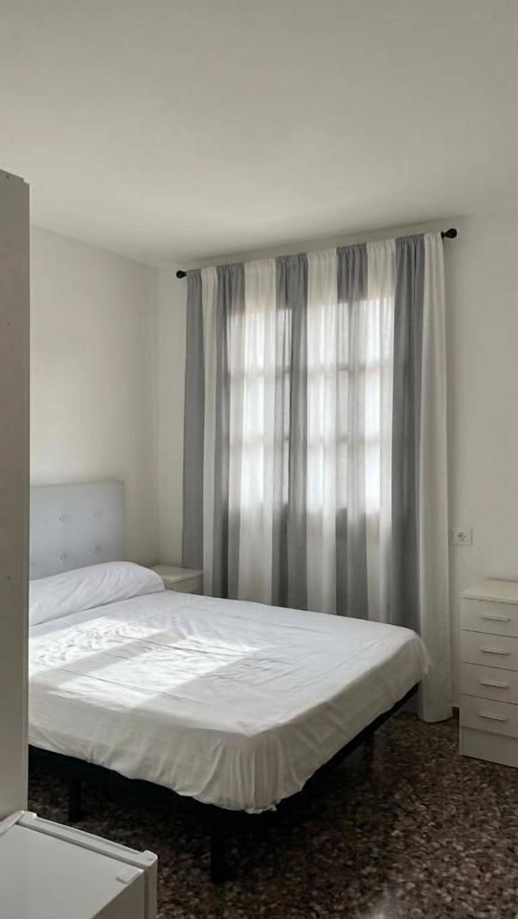 Una cama individual con colchón y almohadas blancos, una mesita de noche, un aparador con cajones y una ventana con cortinas traslúcidas.