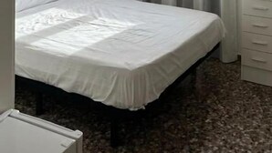 Uma cama de solteiro com colchão e almofadas brancas, uma mesa de cabeceira, um penteado com gavetas e uma janela com cortinas leves.
