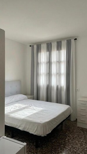 En enkeltseng med hvid madras og puder, et natbord, en kommode med skuffer og et vindue med let gardin.