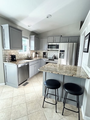 A modern kitchen with grey cabinets, stainless steel appliances, a granite countertop, and two bar stools.