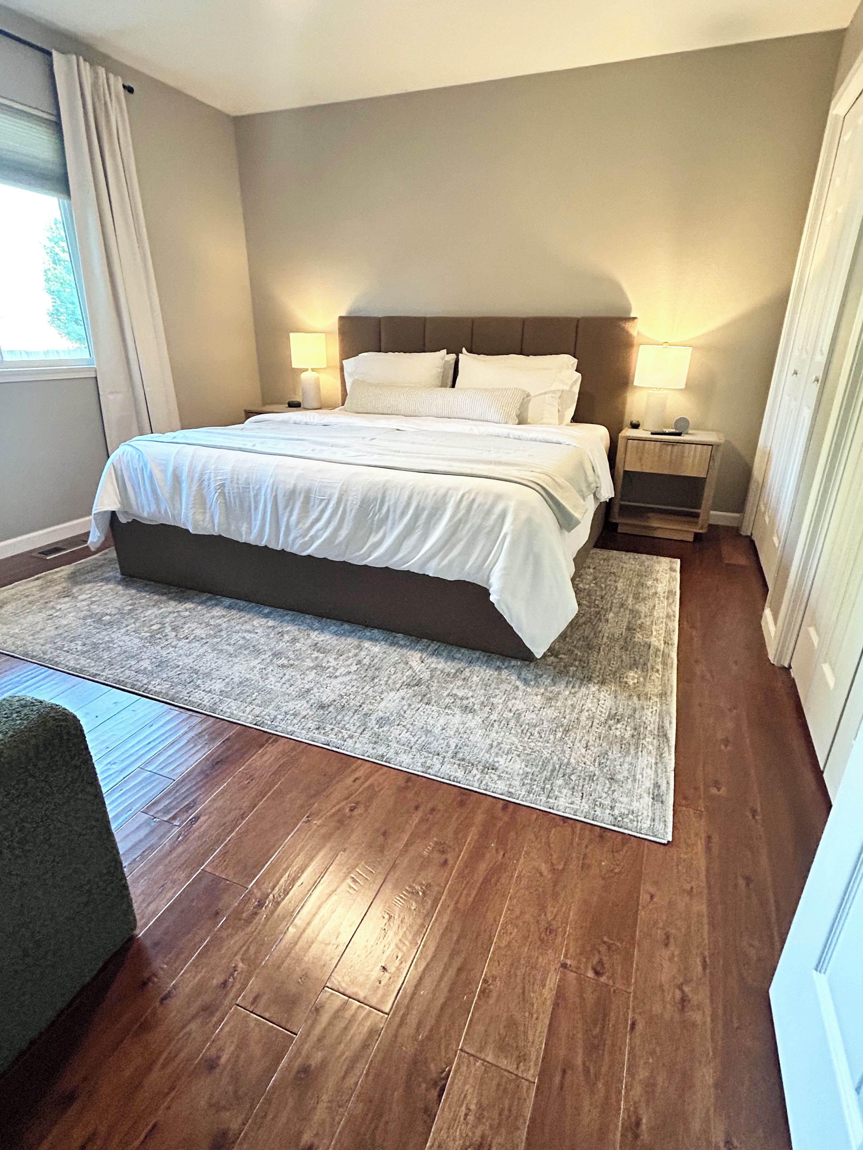 Quarto com uma cama grande, duas mesas de cabeceira, uma mesa de apoio, uma janela com cortinas e uma porta.
