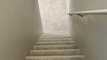 Una escalera alfombrada con barandilla que conduce a una habitación con ventana y una lámpara colgante.