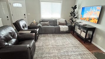 Ein Wohnzimmer mit grauem Sofa, schwarzen Ledersesseln, einem grauen Teppich, einem Fernseher und einem Deckenventilator.