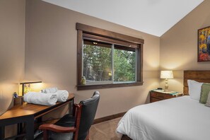 A hotel room with a bed, a desk with a chair, a nightstand, a lamp, and a window with a view of trees. - Chickadee Cabin | Cozy WF Retreat mins to Downtown (Whitefish)