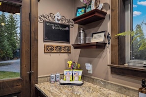 A bar area with a chalkboard sign, a shelf with glasses, and a window with a view of trees. - Chickadee Cabin | Cozy WF Retreat mins to Downtown (Whitefish)