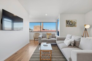 A modern living room with a sofa, a coffee table, and a floor lamp. There is a window with a view of buildings and a chair. - Warwick 2 – Stylish Hobart Retreat (North Hobart)