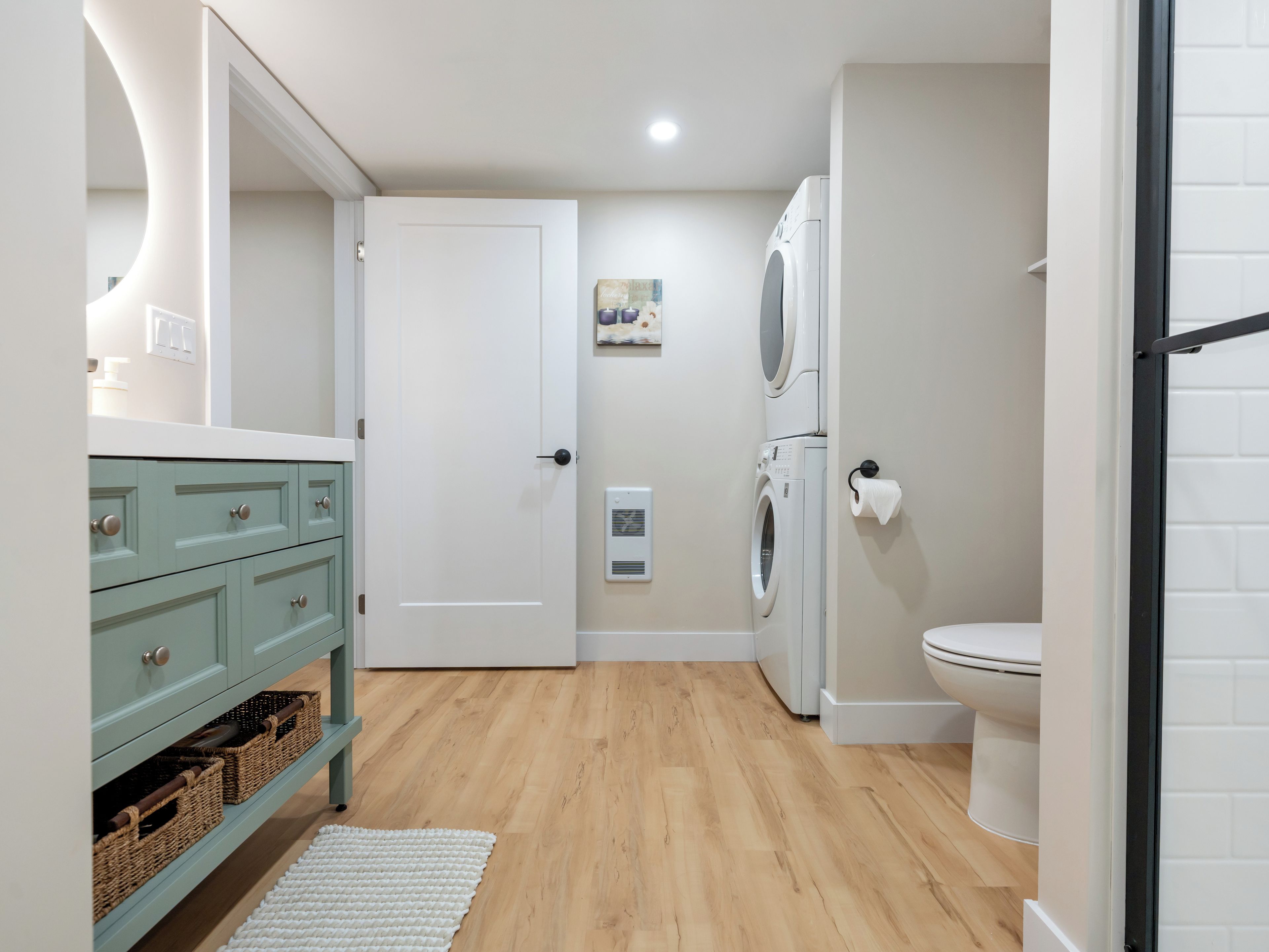 Une salle de bain avec des toilettes blanches, un lave-linge, un meuble-lavabo turquoise avec des tiroirs, un tapis blanc et une porte donnant sur une autre pièce.