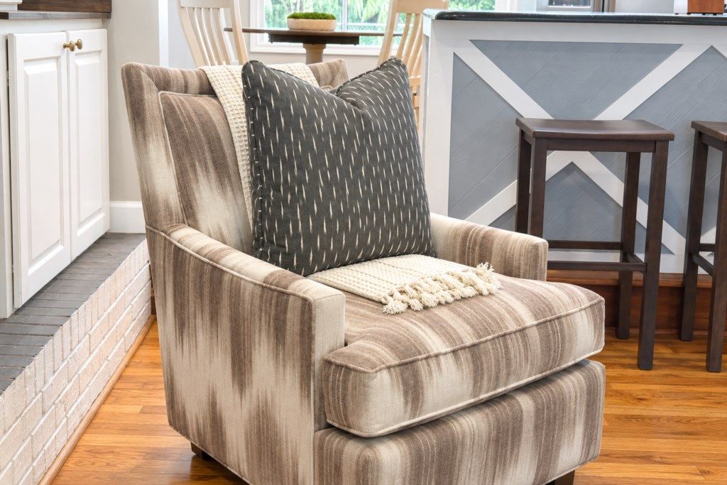 A corner armchair with a striped fabric, a throw blanket, and a patterned pillow in a room with wooden flooring, a kitchen counter, and a chair.