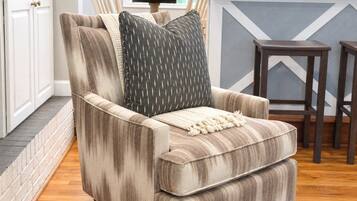 A corner armchair with a striped fabric, a throw blanket, and a patterned pillow in a room with wooden flooring, a kitchen counter, and a chair.