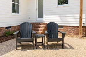 Due poltrone Adirondack e un tavolino davanti a una casa con rivestimento bianco e fondamenta in mattoni.