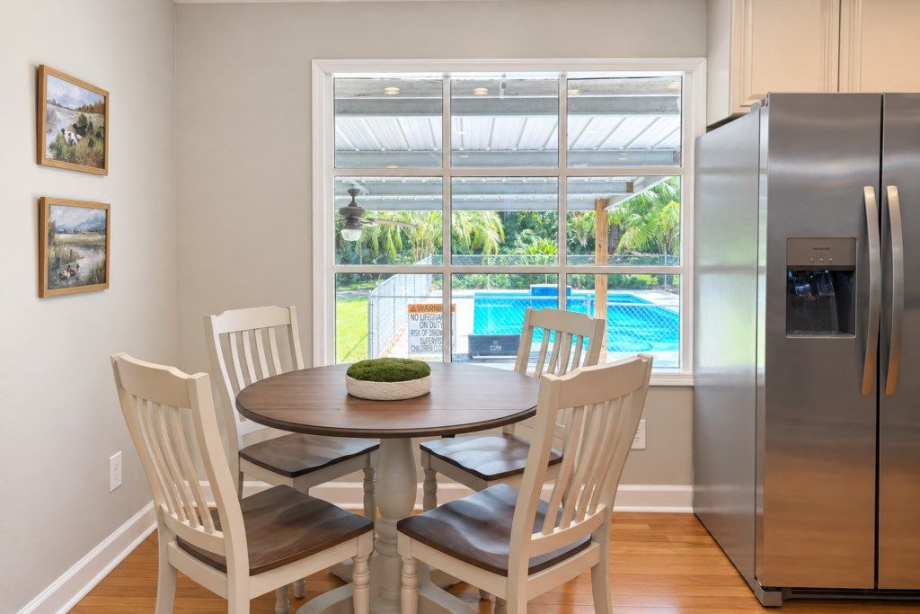 A dining area with a round table and four chairs, a refrigerator, and a large window with a view of a swimming pool and greenery.