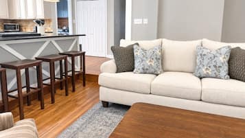 A living room with a sofa, patterned cushions, a wooden coffee table, and a kitchen area in the background.
