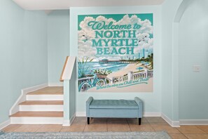 Un escalier menant à une pièce dont un mur est décoré d’une fresque représentant North Myrtle Beach, avec un banc et un mur de couleur bleu clair.