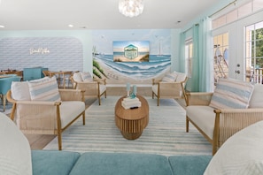 A beach-themed living room with wicker furniture, a central coffee table, and a mural of North Myrtle Beach. - Beach Retreat-Sleeps 22-Outdoor Oasis-Golf Sim-Gym (North Myrtle Beach)