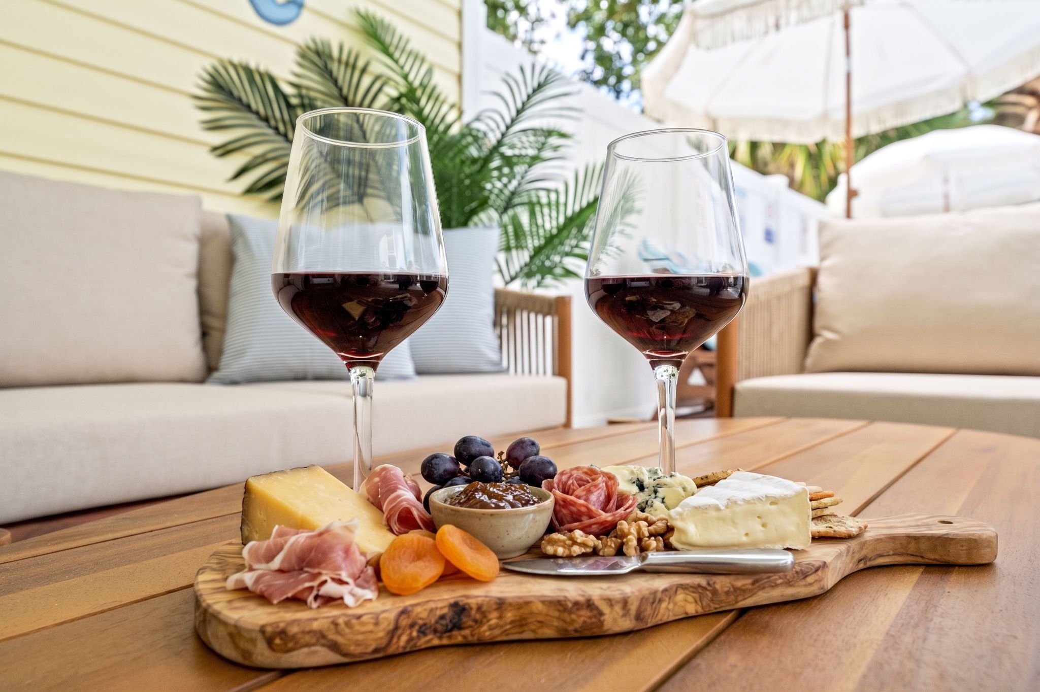 Une table en bois, une planche à fromages, deux verres de vin rouge et un coin salon extérieur accueillant.