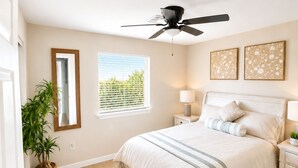 Une chambre à coucher comprenant un lit, des tables de chevet, un ventilateur de plafond, une fenêtre avec des stores et une plante.