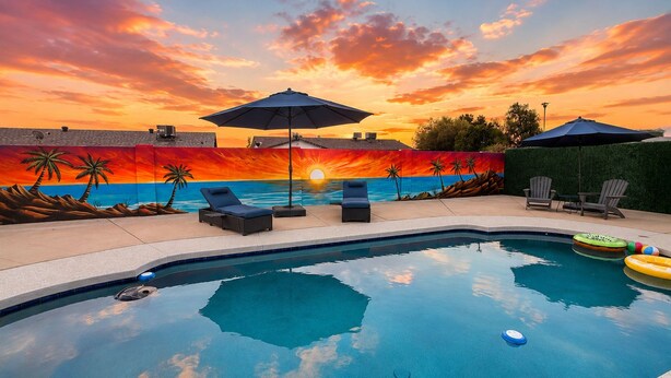 Une piscine entourée de chaises longues et de parasols, avec une fresque représentant un coucher de soleil en arrière-plan.