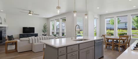 A modern kitchen with a central island, a dining area with wooden chairs, and a living room with a sofa and television.