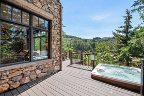 A wooden deck with a hot tub and stone walls, overlooking a forested area. - Exclusive Deer Valley 5BR | Hot Tub, Game Room, Views & Ski Shuttle (Park City)