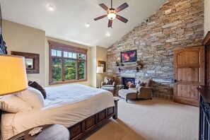A bedroom with a large bed, a stone fireplace, a ceiling fan, and a window with a view of greenery. - Exclusive Deer Valley 5BR | Hot Tub, Game Room, Views & Ski Shuttle (Park City)