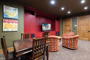 Una habitación con una mesa de comedor de madera, dos sillas, un televisor sobre un mueble rojo y dos otomanes estampados.