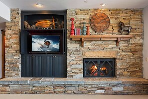 A stone fireplace with a television mounted above it, a clock on the wall, and a mounted shelf with decorative items. - Exclusive Deer Valley 5BR | Hot Tub, Game Room, Views & Ski Shuttle (Park City)