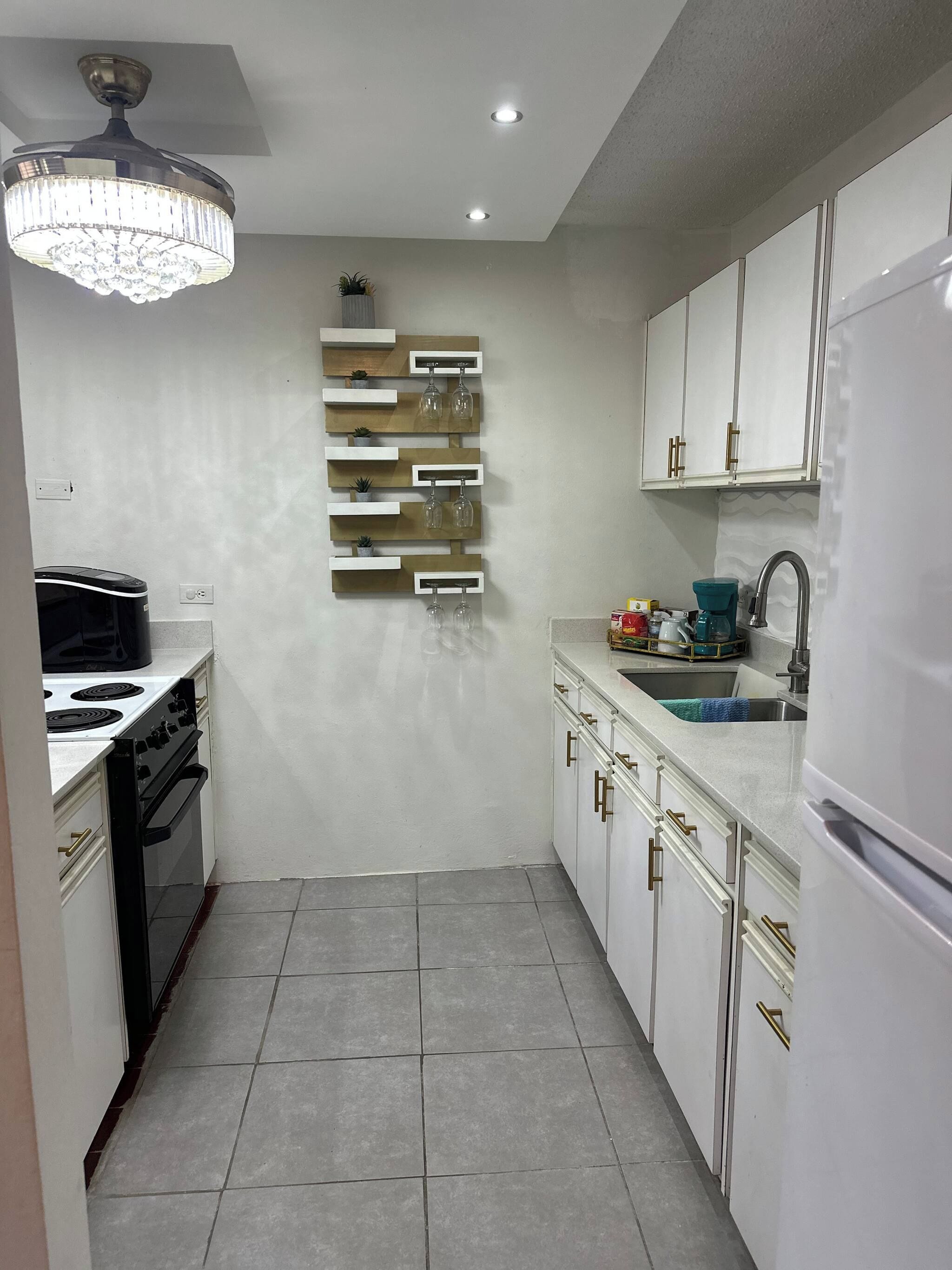 A compact kitchen with white cabinets, a built-in oven, a sink, and a decorative wall-mounted shelf with glasses.