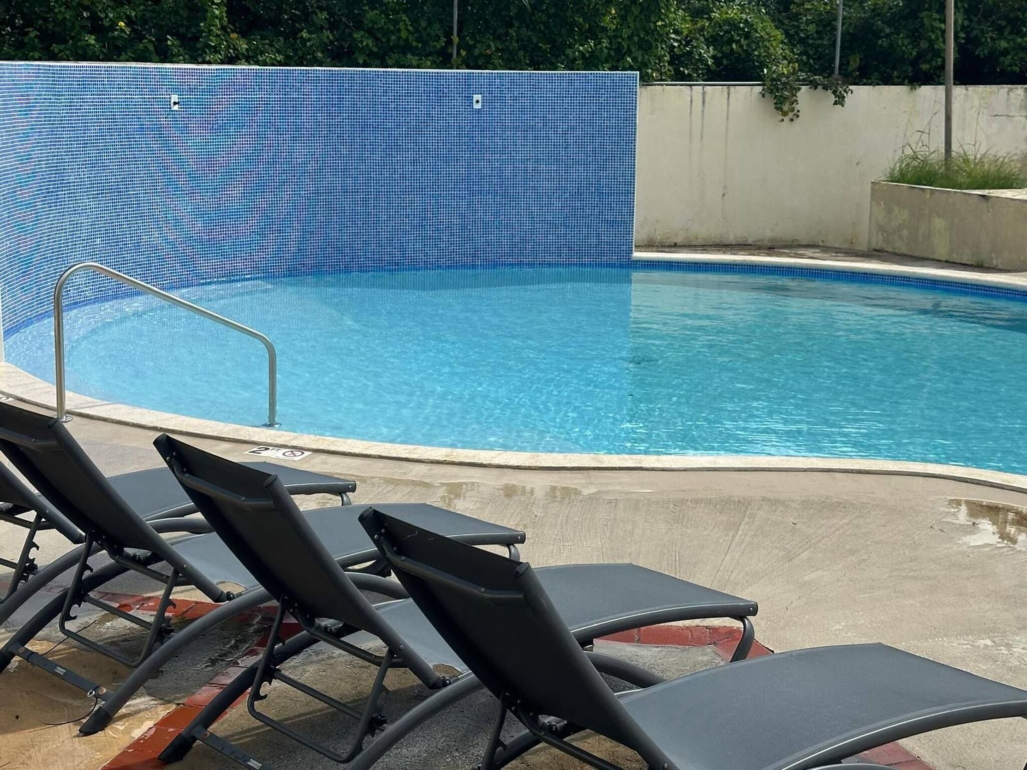 A swimming pool with a blue tiled edge and a metal safety railing. There are black lounge chairs along the poolside.