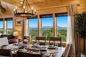 Ein Essbereich mit einem Tisch für vier Personen, große Fenster mit Blick auf eine Berglandschaft und eine Decke mit Holzbalken und hängenden Lampen.
