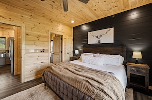 Quarto com teto de madeira, cama com roupa de cama branca, obra de arte pendurada e mesa de cabeceira com abajur.