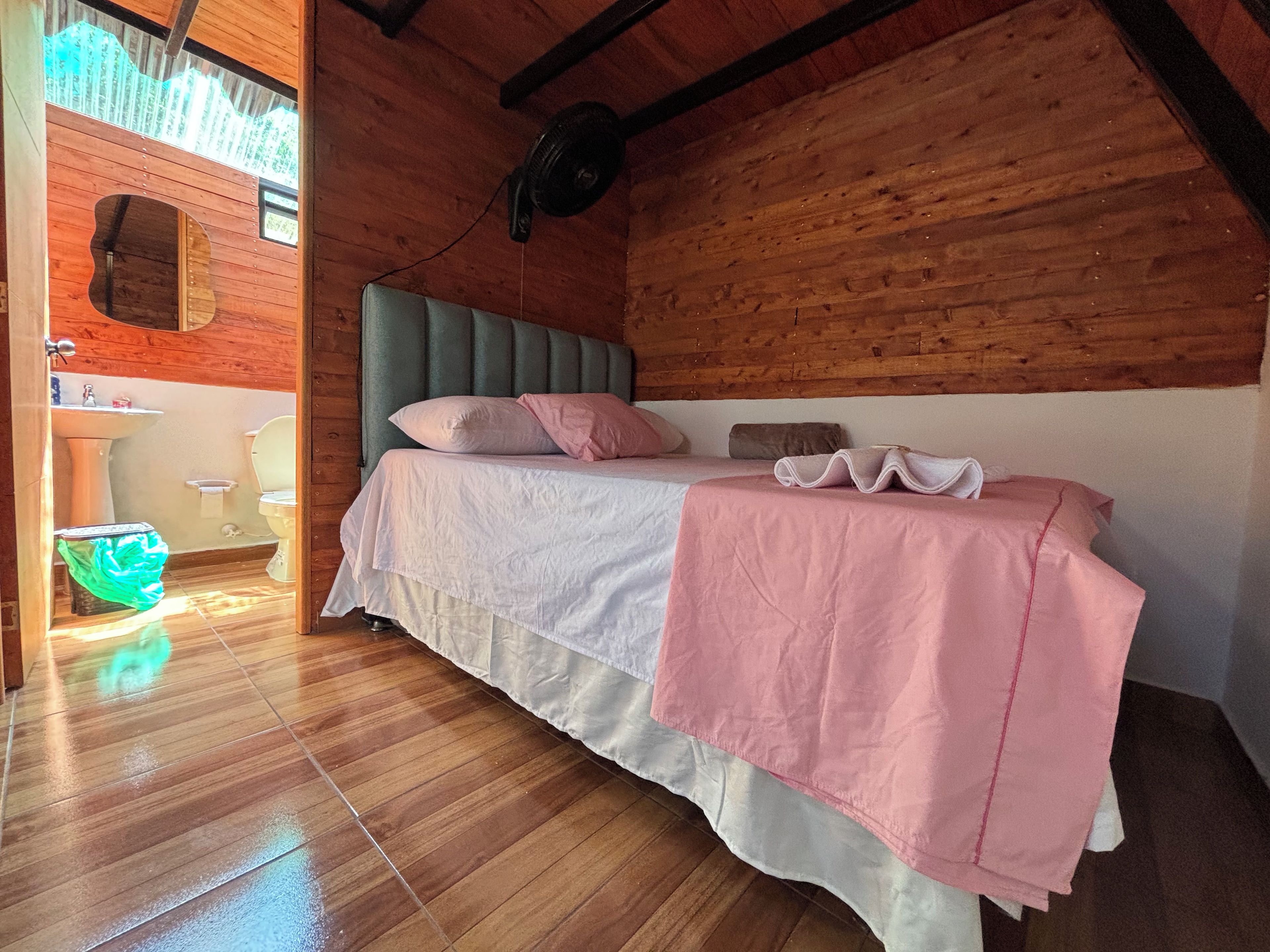 Ein Zimmer mit Holzboden, einem Bett, einem Fenster und einem durch eine geöffnete Tür sichtbaren Badezimmer.