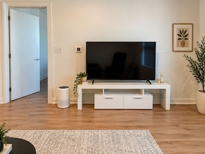 Ein modernes Wohnzimmer mit einem Flachbildfernseher auf einem weißen Standfuß, einer Topfpflanze und einem kleinen Lautsprecher.
