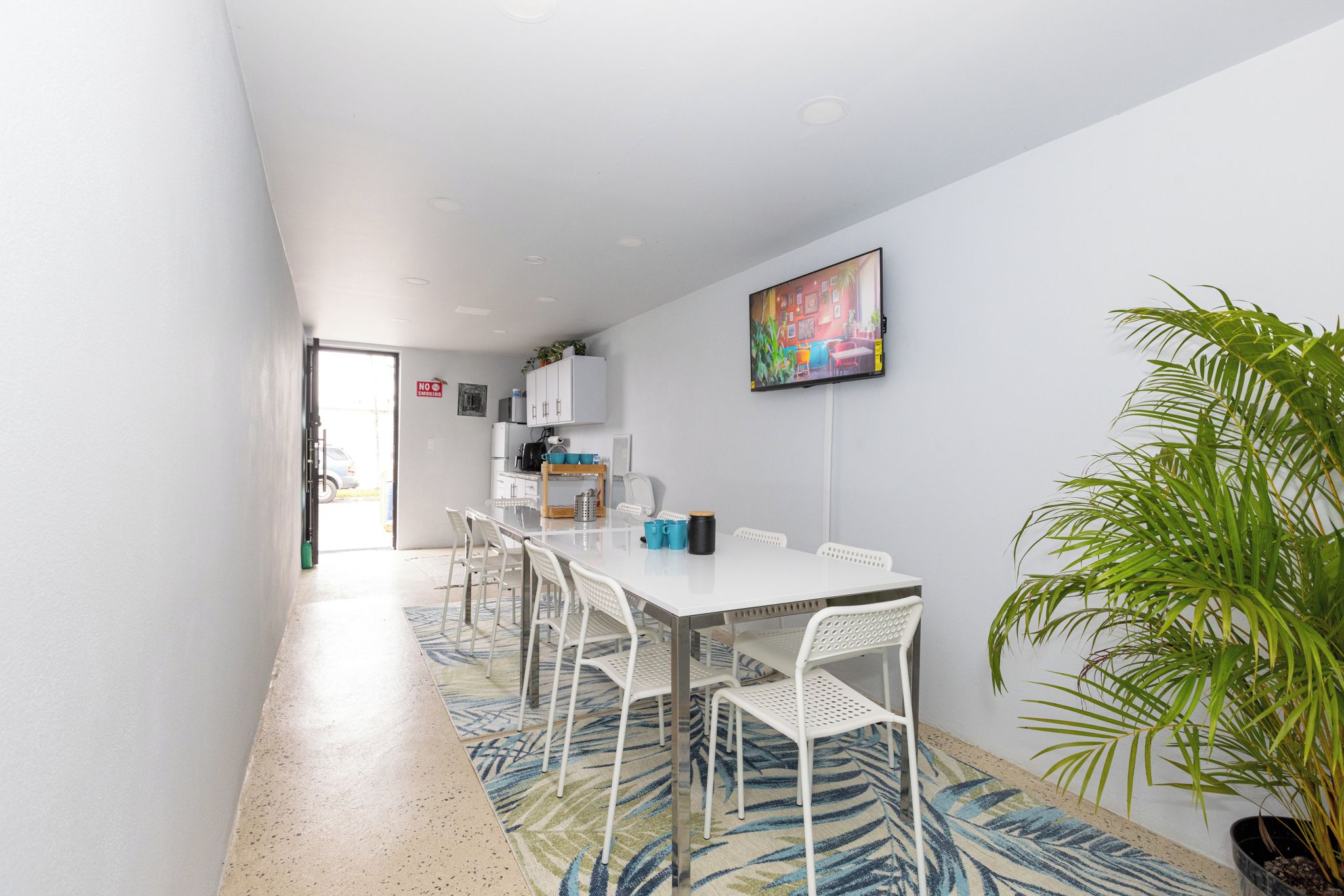 Un comedor moderno con una mesa y sillas blancas, un televisor montado en la pared y una planta en maceta.