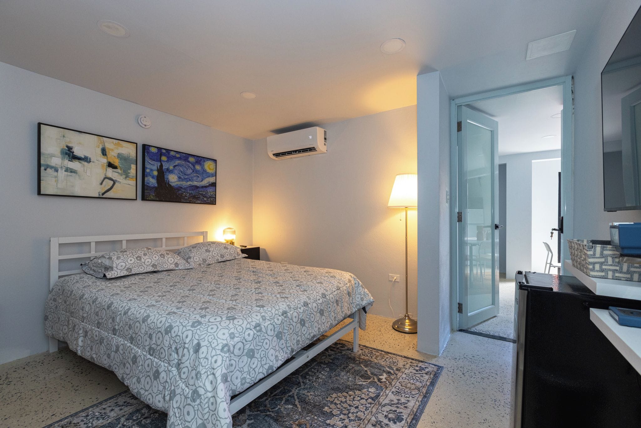 Un dormitorio con cama, lámparas de noche, aire acondicionado instalado en la pared y una puerta que da a otra habitación.