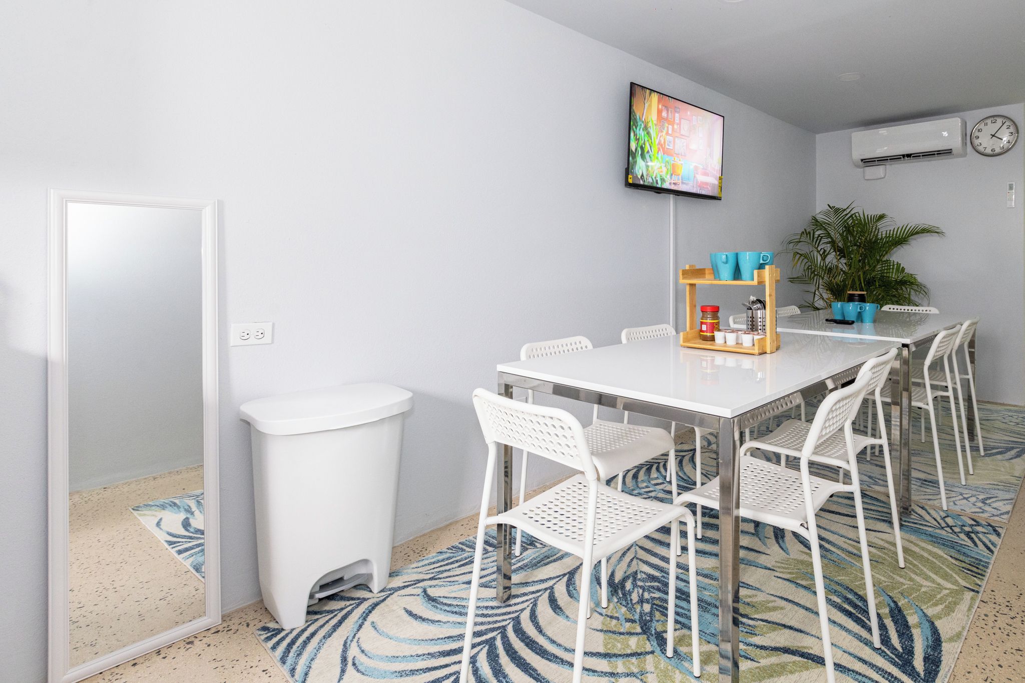 Una habitación moderna con una mesa y sillas blancas, un televisor, una planta en maceta y un reloj en la pared.