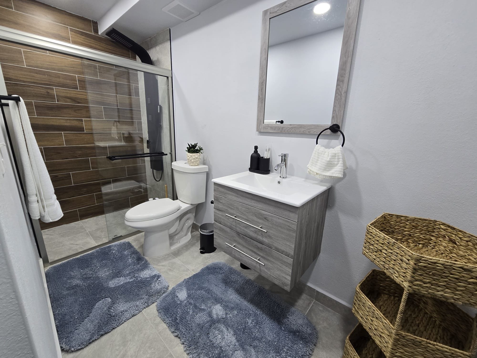 Une salle de bain moderne avec une douche à parois de verre, des toilettes, un meuble-lavabo avec un miroir et deux tapis bleus.