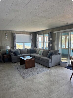 A living room with a grey sectional sofa, a wooden coffee table, and a rug. There are curtains on the windows, and a lamp on a side table. - JUST RIGHT OBX 3-bedroom house in Nags Head on the water with beautiful sunsets (Nags Head)